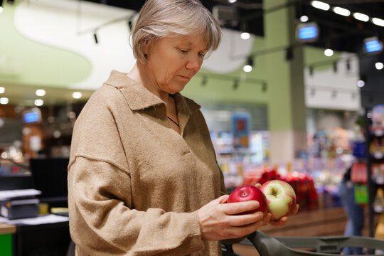 A woman examines two apples in a grocery store, deciding between a red and a green one. The store features bright lighting and shelves full of products