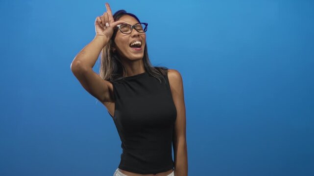 Young thai woman with glasses sticks out tongue and makes l hand to forehead in blue studio; playful mischief.