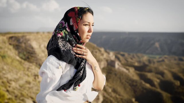 Young thoughtful woman wearing a traditional floral headscarf standing outdoors. She is peacefully contemplating the hilly landscape on a windy day