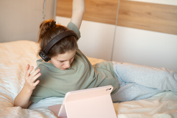 A teenage girl lies on her bed wearing headphones, using a tablet for learning or communication, illustrating digital leisure, online interaction, and a cozy home environment
