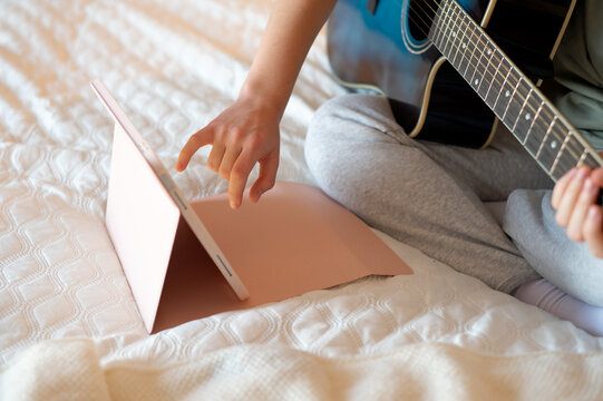 A teenage girl sits with a guitar, learning to play using a tablet program, illustrating online music learning, digital technology, and engaging leisure activities at home
