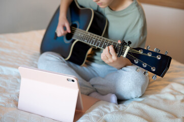 A teenage girl sits with a guitar, learning to play using a tablet program, illustrating online music learning, digital technology, and engaging leisure activities at home