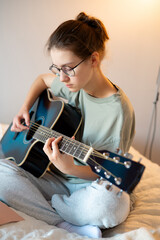 A teenage girl sits with a guitar, practicing playing, showing the learning process, concentration, engagement with music, and creative activity in a cozy home environment