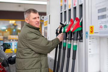 A young man at a gas station picks up a fuel nozzle to fill his car, showing everyday refueling, urban infrastructure, and the interaction between people and modern transportation vehicles