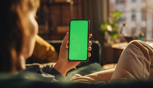 Vertical Screen: Feminine Hand Holding on Smartphone with Green Screen Mock Up Display. Female Resting at Home and Checking Social Media on Mobile Device. Close Up Over the Shoulder Footage