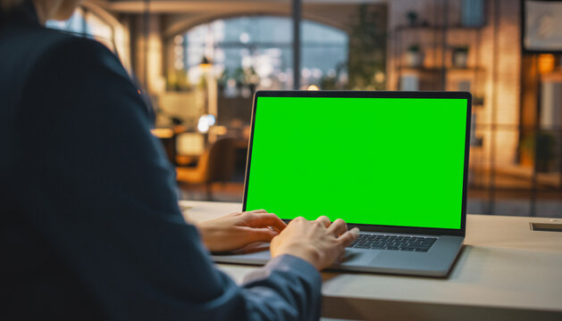 Over the shoulder shot of a business woman working in office interior on pc on desk, looking at green screen. Office person using laptop computer with laptop green screen