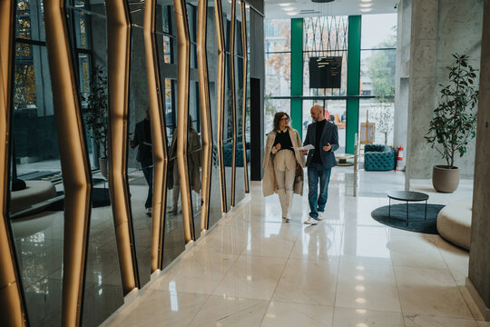 Two professionals stroll through a sleek lobby, sharing papers in a business discussion.