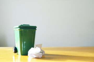 A small green waste bin with crumpled paper on a wooden table stands in natural light. Highlighting sustainable living, eco habits, minimal waste, recycling culture, and environmental awareness. 