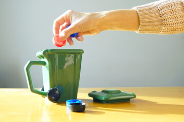 Hand holding plastic bottle caps above green garbage bin. Symbolizes interactive sorting exercise, waste type identification, environmental game-based learning, and hands-on sustainability education.