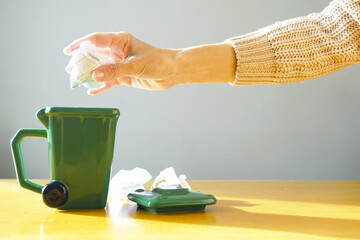 Person crumpling paper above a small green trash container with a detached lid nearby. Embodies recycling habits formation, environmental mindfulness, and waste sorting methodology demonstration.