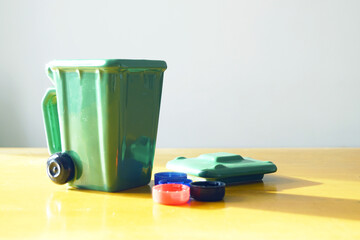 Green small waste bin with colorful bottle caps arranged as sorting demonstration. Concepts of plastic garbage separation, micro-plastic pollution awareness, and waste segregation education.