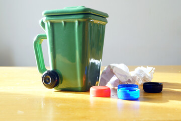 Small green trash bin surrounded by bottle caps and crumpled papers. Symbolizes household waste sorting, proper garbage disposal education, environmental awareness at home, developing recycling habits