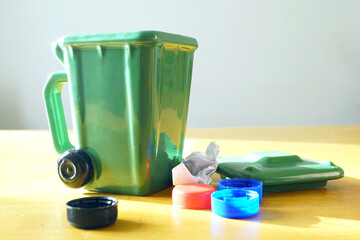 Miniature wheeled garbage container with colorful plastic caps and paper waste beside it. Represents sustainable living practices, zero-waste lifestyle promotion, and domestic waste management.
