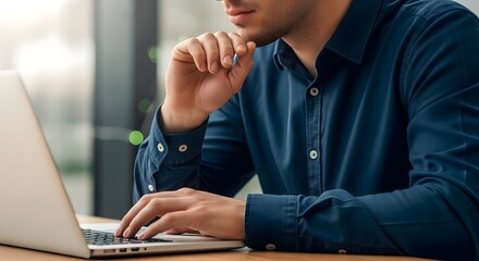 A man in a blue shirt is typing on a laptop while resting his chin on his hand in a well lit room area