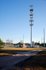 Cell Tower Antenna Mast Rises Against Clear Blue Sky With Wires and Infrastructure. Tall telecom tower with numerous antennas stands against a vivid blue sky; wires and cables run at its base, highlig