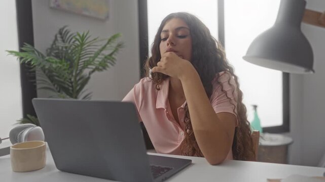 Woman typing on laptop with hand on chin at studio desk, headphones and coffee mug near keyboard; concentration study.