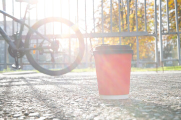 Coffee in a red cup against a backdrop of a bicycle and a fence outside, lifestyle concept with sun rays and glare.	