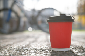 A red coffee cup sits on the asphalt against a backdrop of a bicycle and a fence outside, selective focus, viewed from below. Lifestyle concept with sunbeams and glare.