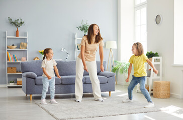 Fototapeta premium Young happy Caucasian mother having fun and dancing with her children in the living room at home. Woman parent enjoying spending time with kids. Family leisure and mothers day concept.