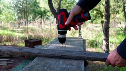 A man is using an electric cordless drill outdoors to make holes to a wooden plank.