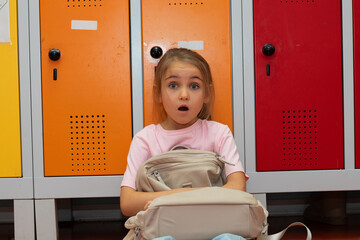 Young girl with surprised expression sitting on the floor in front of colorful school lockers, holding a backpack, capturing a moment of curiosity and wonder in an educational environment