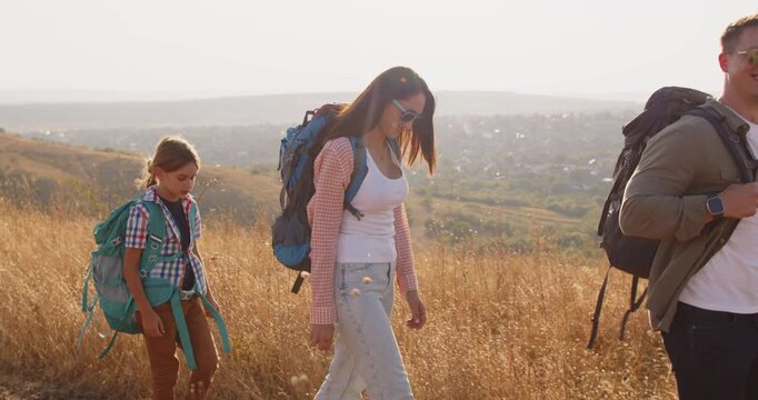 Family hiking with backpack in open nature. They traverse a grassy slope at golden hour, with children exploring trail and enjoying travel together. Concept: family hiking adventure together.
