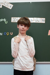 Young boy with brown hair wearing a light shirt and backpack stands in front of a chalkboard displaying numbers and letters, showcasing a classroom environment and educational setting