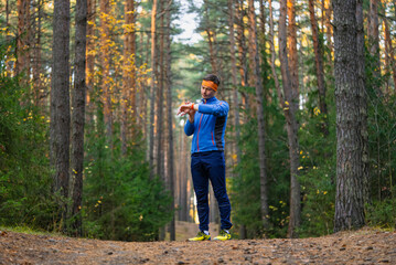 Fototapeta premium Man training on an autumn forest trail checks his smartwatch, standing amid tall pine trees and sunlight while tracking workout data and focusing on fitness goals and performance