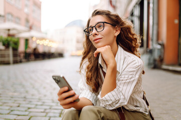 A portrait of a stylish woman with a phone stands in the city's central square at sunset. A beautiful woman enjoys a stroll on a sunny day. Concept of style, fashion, and blogging.