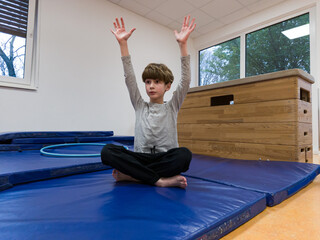 Young boy practicing yoga poses on blue mats in a bright gym, demonstrating balance and focus while surrounded by exercise equipment and natural light