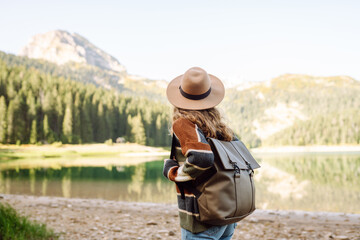 A young woman hiker wearing a hat and carrying a backpack on a hiking trail overlooking a mountain lake. The hiker enjoys the mountain scenery and feels a sense of freedom. Concept: adventure, nature.