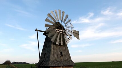 Traditional windmill rotating in green field under blue sky. Blades turning in rural landscape. Documentary footage of countryside wind power and natural scenery.