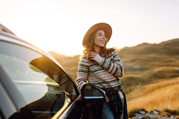 A female traveler stands next to a car on a mountainside on a sunny day. The happy woman enjoys the mountain scenery, feeling the freedom of a road trip. Concept of hiking, freedom.