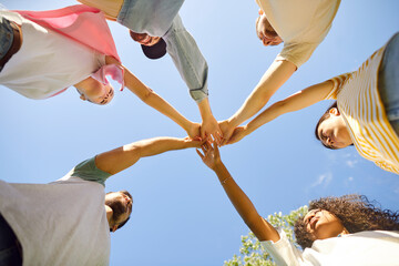 Group of friends stacking hands. Team of young diverse people standing in circle under clear blue summer sky and holding hands together. Bottom view. Friendship and teamwork concept