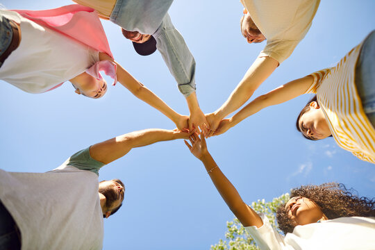 Group of friends stacking hands. Team of young diverse people standing in circle under clear blue summer sky and holding hands together. Bottom view. Friendship and teamwork concept