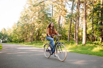 A cheerful woman rides a bicycle along a path in a vibrant green forest. A young woman enjoys a bike ride through a park on a sunny day. Concept: leisure, weekend. Active lifestyle.
