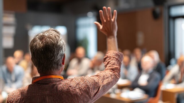A man raising his hand in a conference setting, wearing a patterned shirt and lanyard. He is part of a business meeting and he is asking a question to the speaker or leader. - Powered by Adobe