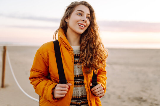 A smiling woman in a bright yellow jacket strolls along a sandy beach at sunset. A female hiker with a backpack enjoys the seascape. Concept of hiking, travel. Lifestyle.