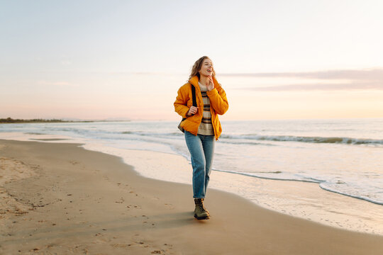 A smiling woman in a bright yellow jacket strolls along a sandy beach at sunset. A female hiker with a backpack enjoys the seascape. Concept of hiking, travel. Lifestyle. - Powered by Adobe