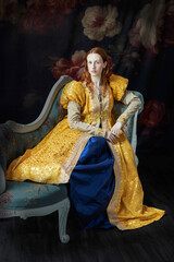 A red-haired woman wearing a historically styled gold gown with puffy sleeves, sitting on an embroidered chaise