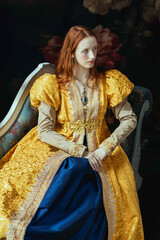 A red-haired woman wearing a historically styled gold gown with puffy sleeves, sitting on an embroidered chaise