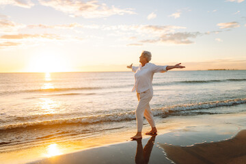 A joyful elderly woman feels free as she strolls along a sandy beach at sunset. Beautiful woman with gray hair enjoys stroll, walking barefoot along seashore. Concepts of nature, aging, retirement.