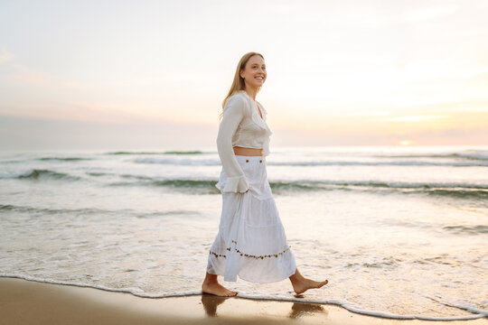 A happy woman in a white dress strolls along the beach at sunset. The young woman enjoys the seascape and feels the freedom of the outdoors. Concept of nature, enjoyment.