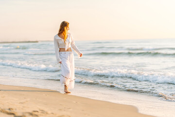 A happy woman in a white dress strolls along the beach at sunset. The young woman enjoys the seascape and feels the freedom of the outdoors. Concept of nature, enjoyment.