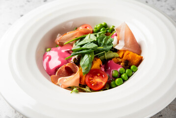 A vibrant gourmet salad with butternut squash, prosciutto, spinach, green peas, cherry tomatoes, and a pink beetroot dressing in a white bowl.