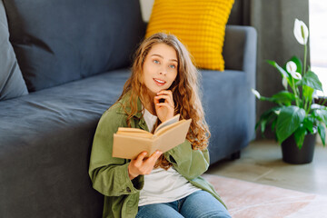 Obraz premium Portrait of a young woman with a book in her hands, relaxing on a sofa in a cozy room. A smiling woman enjoys reading a book indoors on a sunny day. Concepts: education, relaxation.