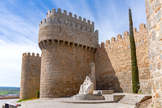 A dramatic view of the Walls of &Aacute;vila with a white stone statue of Saint Teresa in the foreground, seated at the base of a medieval tower beside a tall cypress tree.
