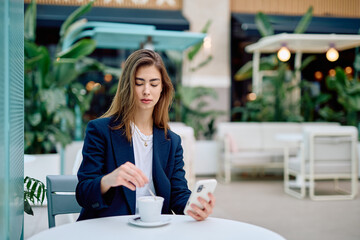 Young woman stirring coffee and checking smartphone at a bustling outdoor cafe. Connecting with remote work and social life