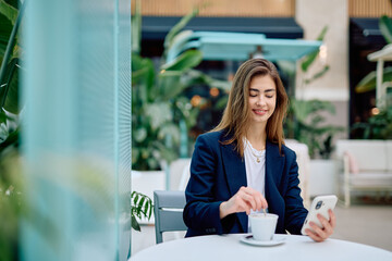Young woman smiling, stirring coffee, and looking at her smartphone at an outdoor cafe. She is engaged with technology