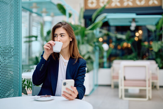 Young woman in business casual attire drinking coffee at a trendy cafe, holding a mobile phone, taking a moment to relax during her day